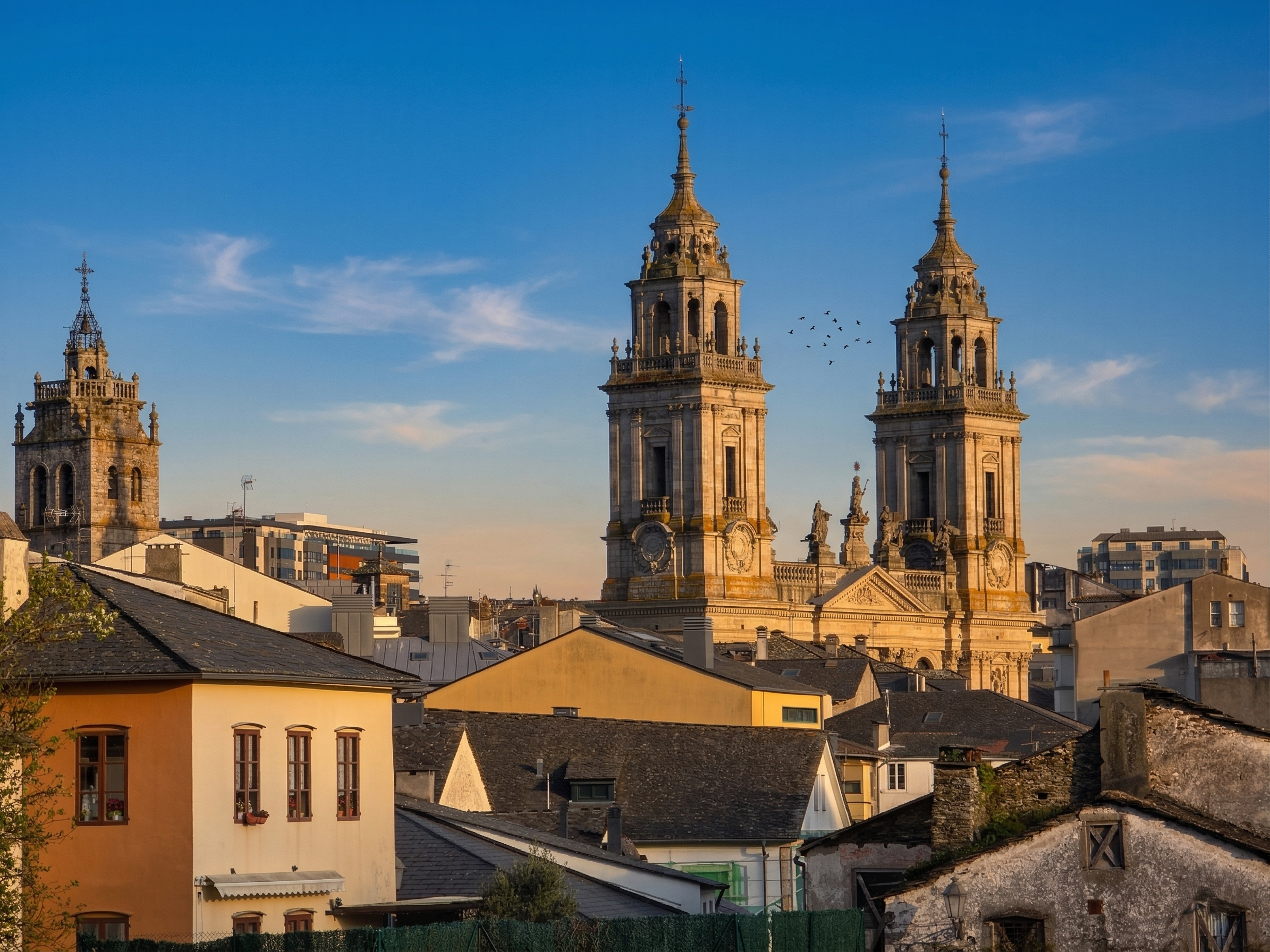 Vista de las torres barrocas de la Catedral de Santa María de Lugo elevándose sobre los tejados tradicionales del casco histórico al atardecer. Un primer plano de edificios antiguos de piedra y tejados de pizarra. Imagen que ilustra la arquitectura histórica de Lugo y la ubicación de los alojamientos gestionados por Urbe Lucus.